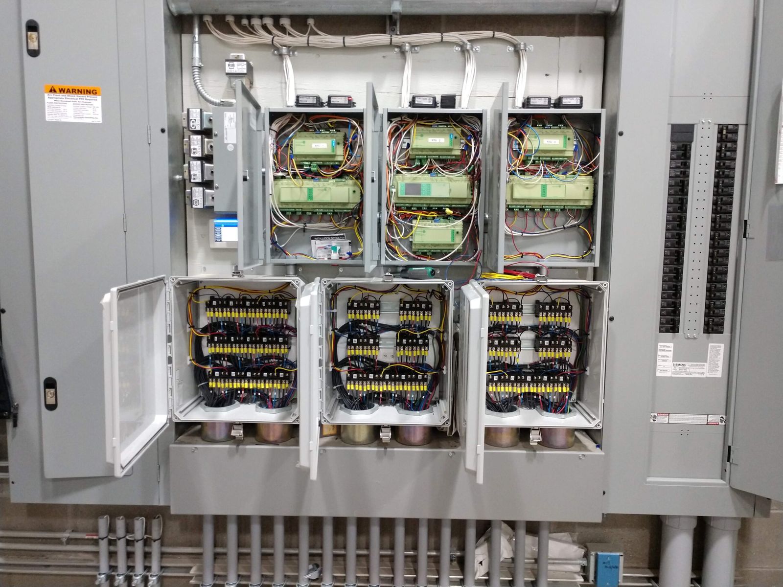 Open control cabinets with three rooftop unit controllers above rows of relays for the floor heating circuits